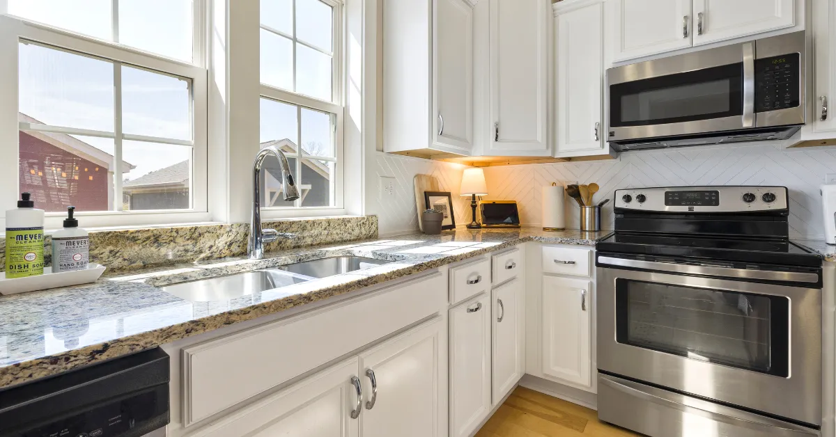 Bright kitchen with white cabinets, granite countertops, a double sink, and stainless steel appliances. Sunlight streams through large windows, creating a welcoming atmosphere.