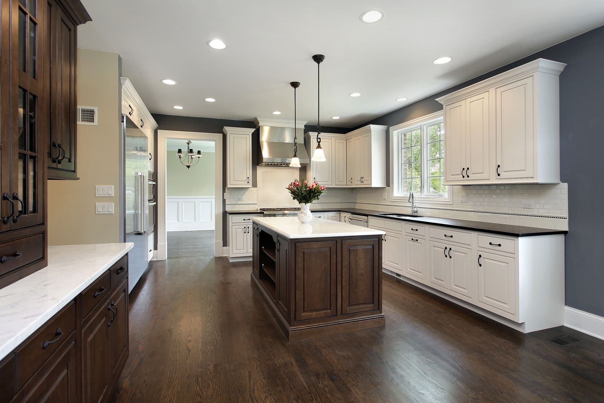 Spacious modern kitchen with dark hardwood floors, white cabinetry, and a central island with a dark wood finish. Large windows let in natural light.