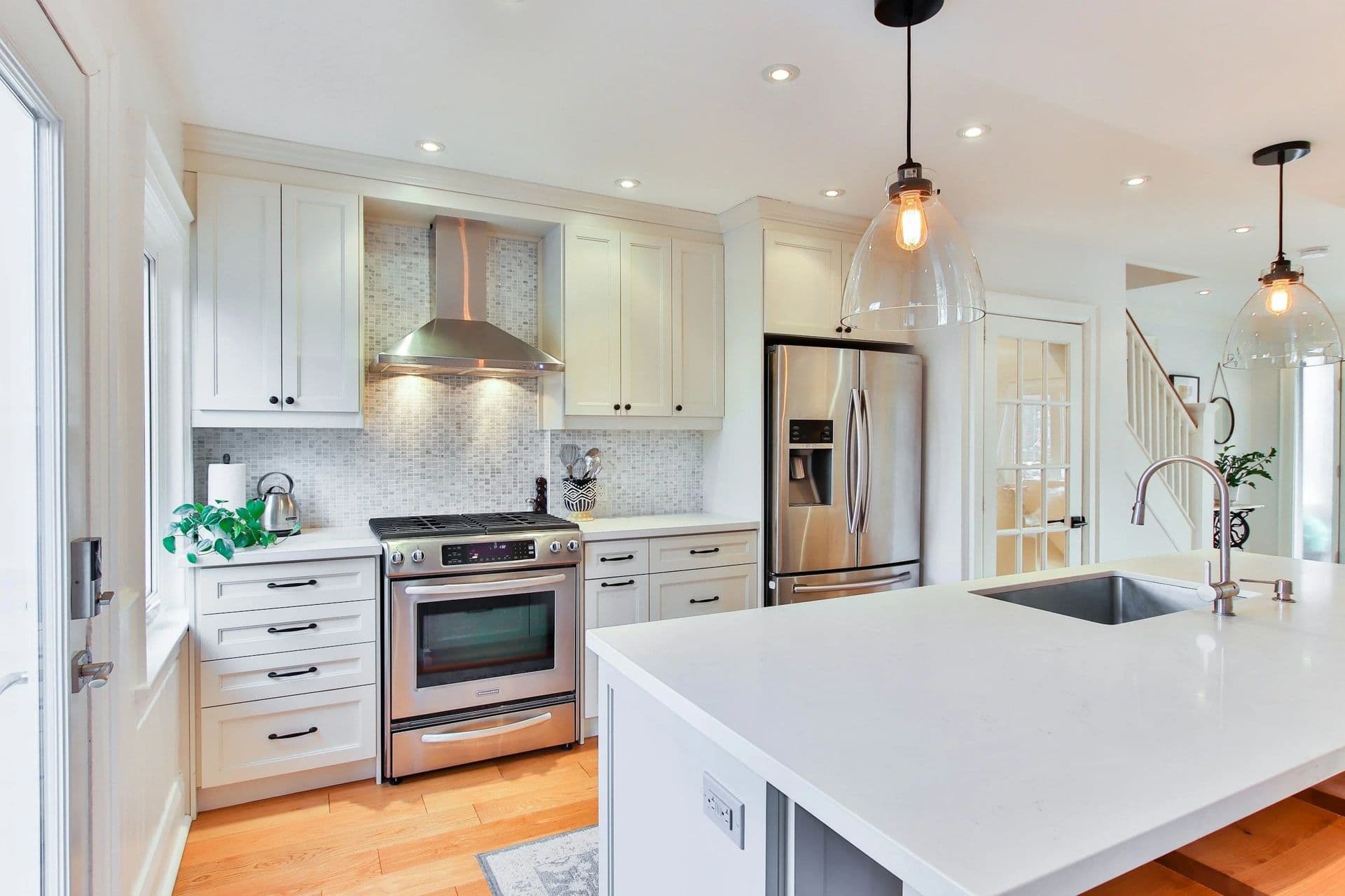 Modern kitchen with white cabinets, stainless steel appliances, a central island with a sink, pendant lights, and wooden flooring, conveying a bright, clean atmosphere.