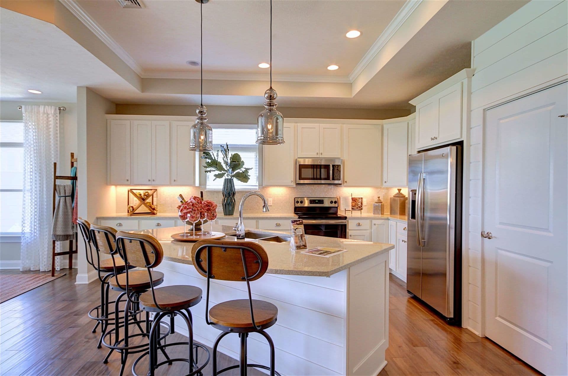 Modern kitchen with wooden floors, white cabinets, and a central island. Three bar stools, pendant lights, and stainless steel appliances add elegance.