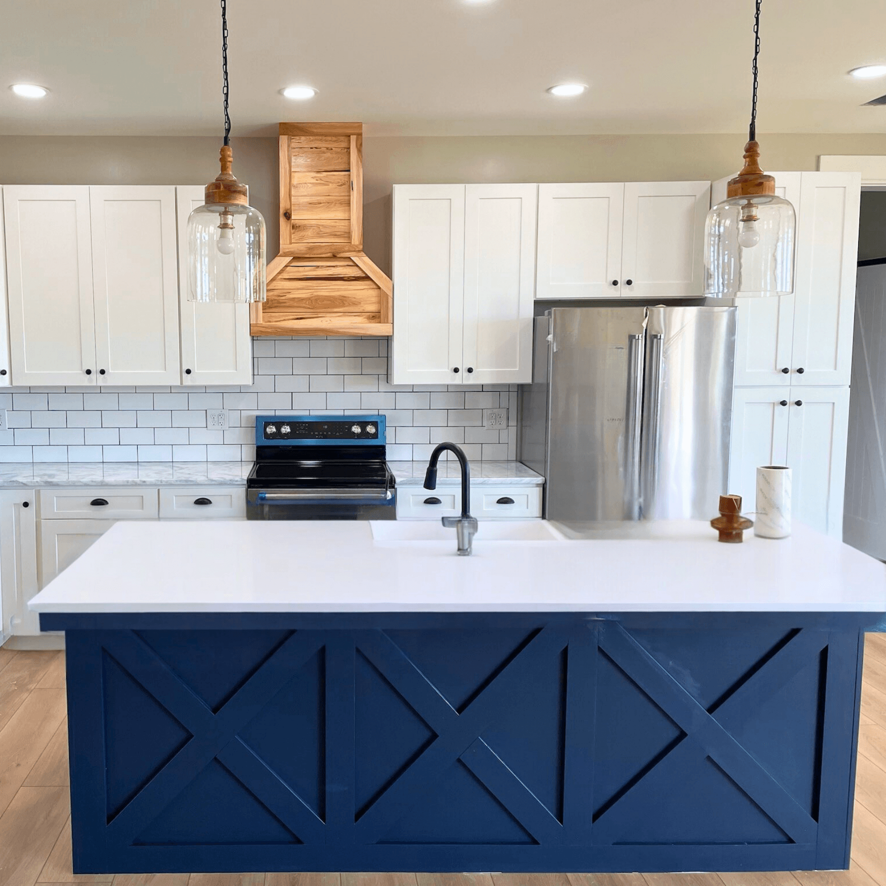 Modern kitchen with white cabinets, subway tile backsplash, and wood range hood. A blue island with white countertop centers the space, under pendant lights.