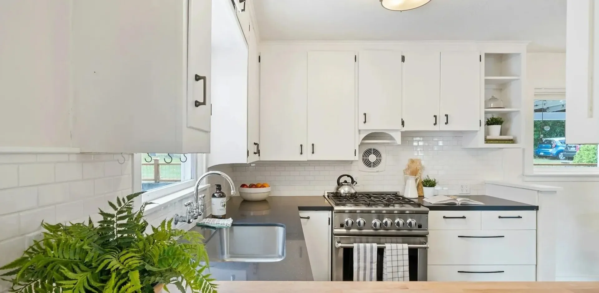 Bright kitchen with white cabinets, stainless steel stove, and a lively green fern on the counter. A cozy and fresh ambiance prevails.
